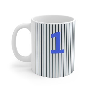 Könnte beinhalten: Weiße Keramiktasse mit einem schwarz-weißen Streifenmuster und der blauen Zahl "1" auf der Vorderseite.