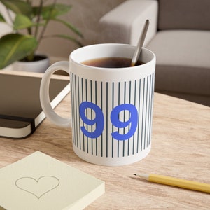 Könnte beinhalten: Eine weiße Keramiktasse mit einem schwarz-weißen Streifenmuster und der Zahl 99 in Blau. Die Tasse ist mit Kaffee gefüllt und hat einen silbernen Löffel darin.