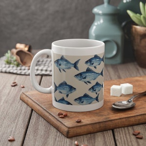 Op de afbeelding: Witte keramische mok met een patroon van blauwe vissen op een beige achtergrond. De mok heeft een witte handgreep.
