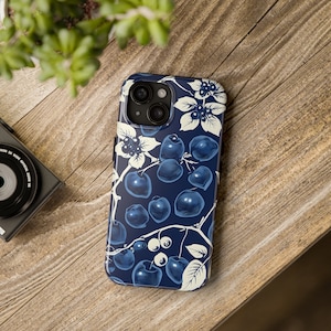 May include: A navy blue phone case featuring a botanical design of blueberries and white floral accents. The case has a glossy finish and is set on a wooden surface. The design includes detailed illustrations of berries and leaves.