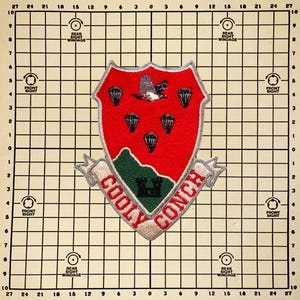 May include: Embroidered patch featuring a red shield with a white and gray bird, parachutes, and a green landscape with a castle. The text "COOLY CONCH" is displayed on a banner below the shield.