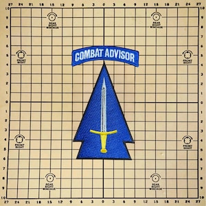Pode incluir: Um patch bordado azul com um desenho de espada e as palavras "COMBAT ADVISOR" em uma faixa curva. O patch está sobre um fundo quadriculado com indicações de ajuste de mira.