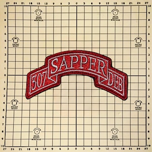 May include: A red and white embroidered patch with the text "507 SAPPER DEB" in white lettering. The patch has a curved banner design and is set against a grid background.