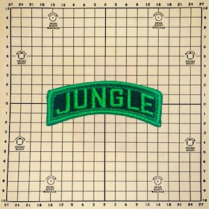 May include: A curved, embroidered patch featuring the word JUNGLE in bold, dark green letters on a black background with a bright green border. The patch is positioned on a grid background with measurements ranging from 0 to 27 inches.