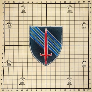 May include: A shield-shaped embroidered patch featuring a black background with a central vertical red sword. Diagonal stripes in blue and green extend from the upper left to the right. The patch has a grey border and is centered on a grid.
