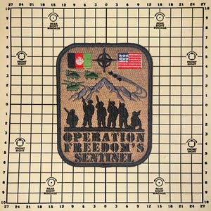 May include: A tan and brown patch with the text "OPERATION FREEDOM'S SENTINEL". The patch features silhouettes of soldiers, flags, mountains, and military aircraft. The flags include Afghanistan and the United States.