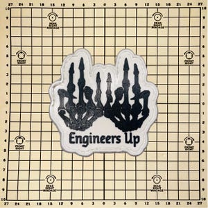May include: A patch featuring a black and white skeletal hand gesture with the text "Engineers Up" in a bold font. The patch has a white border and is set against a grid background.