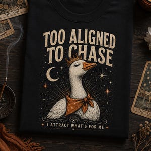 Pode incluir: Uma t-shirt preta com o gráfico de um ganso branco a usar uma coroa e um lenço laranja. O texto "TOO ALIGNED TO CHASE" e "I ATTRACT WHAT'S FOR ME" aparece acima e abaixo do ganso, rodeado por estrelas e uma lua crescente.