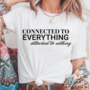 May include: White t-shirt with black text that reads "CONNECTED TO EVERYTHING attached to nothing". The model is wearing the shirt with a brown belt and blue jeans.
