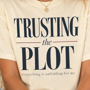 May include: A cream-coloured short-sleeve t-shirt featuring the dark blue text TRUSTING the PLOT and the smaller text everything is unfolding for me. The shirt has a crew neckline and is worn with blue denim jeans and a gold pendant necklace.