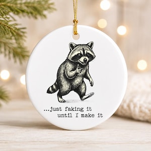 Könnte beinhalten: Ein rundes, weißes Keramikornament mit einer schwarz-weißen Illustration eines Waschbären, der geht und nach vorne zeigt. Unter dem Waschbären steht der Text: ...just faking it until I make it. Das Ornament hängt an einer dünnen goldenen Schnur.