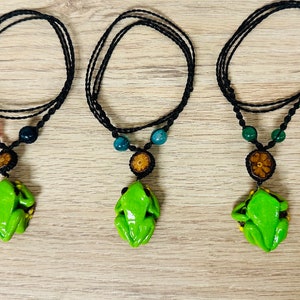 Puede incluir: Tres collares hechos a mano con un colgante de rana verde brillante. Cada collar tiene un cordón negro, cuentas de colores y una pequeña pieza de acento redonda y marrón. Los colgantes de rana tienen detalles amarillos.