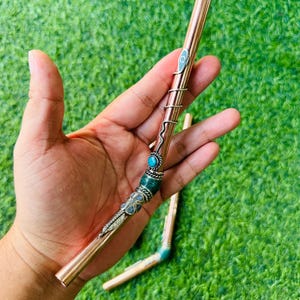 Puede incluir: Una pajita de metal color cobre con detalles decorativos plateados y piedras de color turquesa. La pajita tiene un diseño en espiral y un detalle en forma de pluma. Una segunda pajita es visible en el fondo.