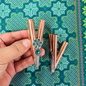 May include: Two copper-coloured metal pipes with decorative silver accents and turquoise-coloured stones. One pipe has a flower design, the other has a leaf design. The pipes are displayed against a patterned green and blue background.