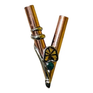Puede incluir: Objeto en forma de V de metal color cobre, con detalles plateados y dorados. El diseño incluye una espiral plateada, un elemento floral negro y dorado y una pequeña piedra verde. Fondo blanco.