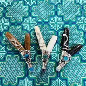 Puede incluir: Cuatro bolígrafos decorativos con detalles plateados, turquesas y otras piedras preciosas. Los barriles de los bolígrafos son de madera, hueso y material negro. Los bolígrafos están colocados sobre una tela estampada verde y azul verdoso.
