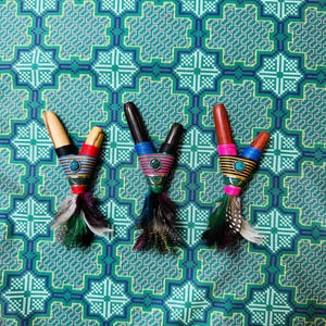Puede incluir: Tres pipas de madera hechas a mano con bandas tejidas coloridas y detalles turquesa. Cada pipa presenta un tono de madera diferente y está adornada con plumas decorativas de varios colores. Las pipas descansan sobre una tela estampada verde y turquesa.