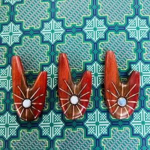 May include: Three V-shaped wooden decorative objects with a sunburst design, central white stone, and copper accents. They rest on a patterned green and blue background. The objects are approximately 10 cm in length.