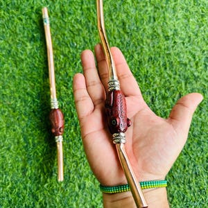 Puede incluir: Dos pajitas de yerba mate de color cobre, una sostenida en una mano. Las pajitas tienen un diseño curvo con un acento decorativo de madera marrón rojizo. Las pajitas están sobre un fondo verde.