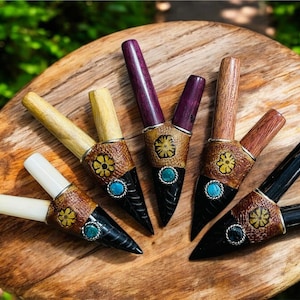 Puede incluir: Colección de seis pipas de madera hechas a mano con bandas de cuero decorativas. Cada pipa presenta una combinación de colores única, incluyendo negro, marrón, morado y crema, con acentos turquesa y amarillos. Las pipas están dispuestas sobre una superficie de madera.