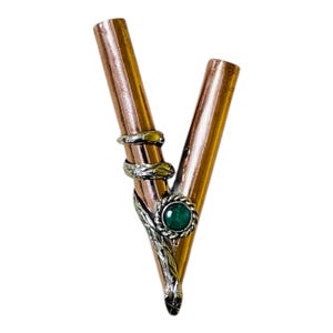 Puede incluir: Tubo de metal color cobre con diseño en forma de V. Está adornado con detalles de metal plateado, incluyendo un diseño en espiral y un engaste circular con una piedra preciosa verde. El diseño es ornamentado.
