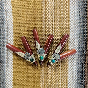 May include: Four wooden pipes with decorative metal accents and turquoise-coloured stones. The pipes are a rich reddish-brown colour and are arranged on a textured, striped fabric background.