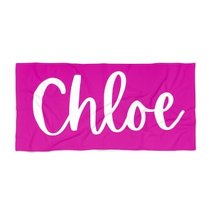 以下が含まれることがあります： 鮮やかなピンク色のビーチタオル。中央に「Chloe」という名前が大きな白い筆記体で書かれています。長方形のタオルは柔らかい生地で作られているようです。