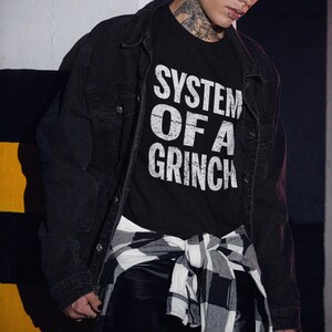 Könnte beinhalten: Ein schwarzes T-Shirt mit weißem Text, der "System of a Grinch" lautet. Das Shirt wird von einer Person mit einer schwarzen Jacke und einem karierten Hemd getragen, das um die Taille gebunden ist.