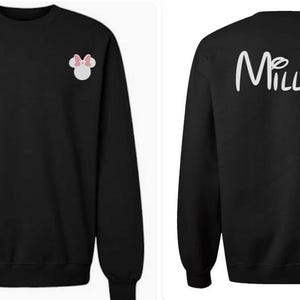 May include: A black long-sleeve sweatshirt featuring a small white mouse head icon with a pink bow on the front left chest and the white text "Millan" in a cursive script font printed across the upper back area of the garment.