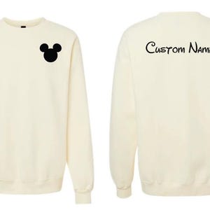 May include: A cream-colored long-sleeve crewneck sweatshirt. The front features a small black silhouette of a mouse head on the left chest. The back displays the text Custom Name in a black script font centered near the top of the garment.