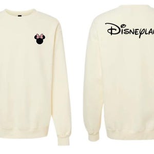 May include: A cream-colored crewneck sweatshirt featuring a small black Minnie Mouse silhouette with a pink bow on the left chest. The back of the sweatshirt displays the word Disneyland printed in a large, black, stylized cursive script font.