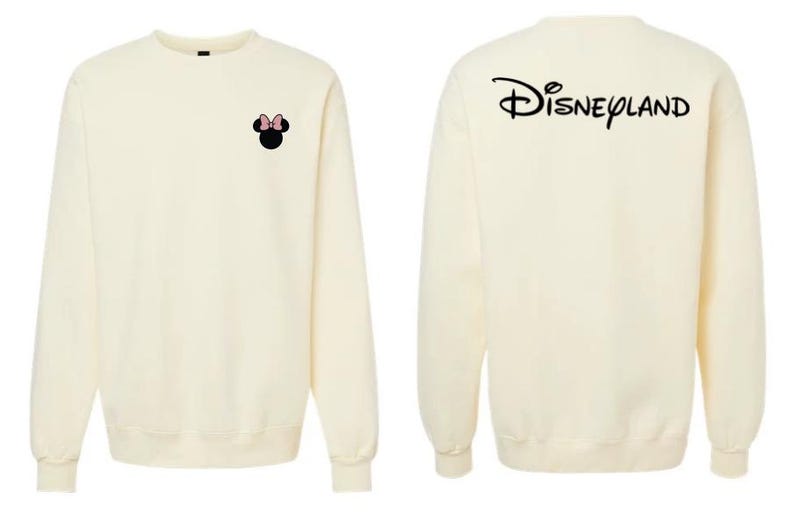 May include: A cream-colored crewneck sweatshirt featuring a small black Minnie Mouse silhouette with a pink bow on the left chest. The back of the sweatshirt displays the word Disneyland printed in a large, black, stylized cursive script font.