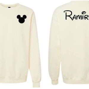 May include: A cream-colored crewneck sweatshirt featuring a black silhouette of a mouse head on the left chest and the text "Ramirez" in a stylized black font across the upper back. The garment has long sleeves and a ribbed collar, cuffs, and hem.