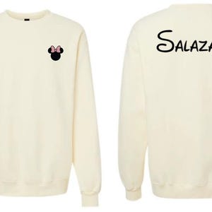 May include: A cream-colored long-sleeve sweatshirt featuring a small black Minnie Mouse head icon with a pink bow on the front left chest and the name Salazar printed in a black stylized script font across the upper back of the garment.