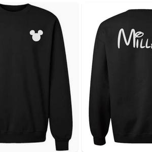 May include: A black long-sleeve crewneck sweatshirt featuring a small white Mickey Mouse head silhouette on the left chest and the word "MILLAN" in a white script font on the back, with the letter "i" dotted by a small Mickey Mouse head.