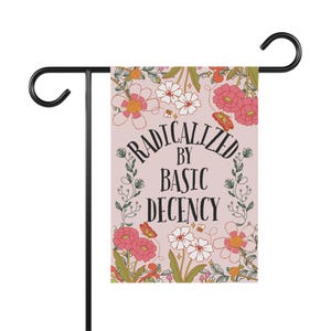 May include: A garden flag with the text "RADICALIZED BY BASIC DECENCY" in black script, surrounded by floral illustrations in pink, white, and orange. The flag is mounted on a black metal stand.