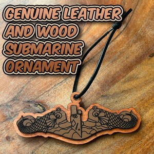 May include: A wooden submarine ornament with a leather inlay, featuring a black cord for hanging. The ornament has the text "GENUINE LEATHER AND WOOD SUBMARINE ORNAMENT" in a brown and black font. The submarine design includes fish and a submarine.