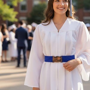 Pode incluir: Uma pessoa vestindo uma beca branca com um cinto azul e uma fivela de madeira. A pessoa est&aacute; sorrindo e usando um chap&eacute;u de formatura. Outras pessoas est&atilde;o ao fundo.