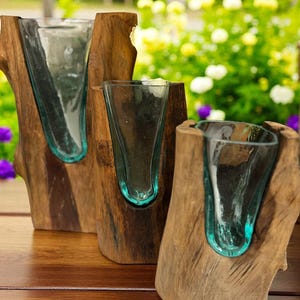 May include: Three rustic wooden vases with teal-colored glass inserts. The vases are made from natural wood, showcasing the grain and texture. The glass inserts are shaped to hold flowers or other decorative items. The background features a blurred view of greenery and flowers.