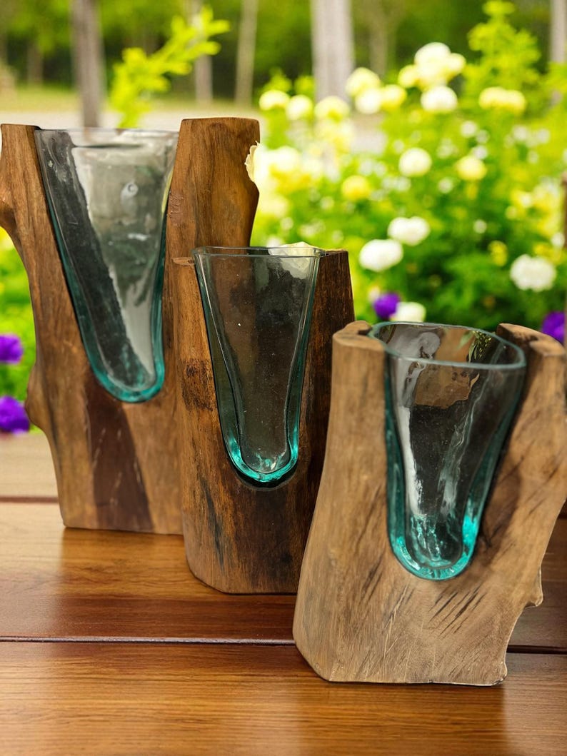 May include: Three rustic wooden vases with teal-colored glass inserts. The vases are made from natural wood, showcasing the grain and texture. The glass inserts are shaped to hold flowers or other decorative items. The background features a blurred view of greenery and flowers.