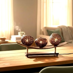 May include: A decorative table lamp featuring three spherical, dark brown, perforated shades arranged in a row on a curved wooden base. The lamp is supported by two vertical posts wrapped in light-colored cord, resting on a rectangular wooden stand.