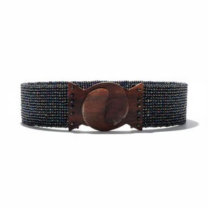 Pode incluir: Um cinto largo com uma faixa de contas em tons de preto e iridescentes. O cinto apresenta uma grande fivela de madeira escura com um design curvo &uacute;nico. A fivela &eacute; presa com pequenos rebites de metal.