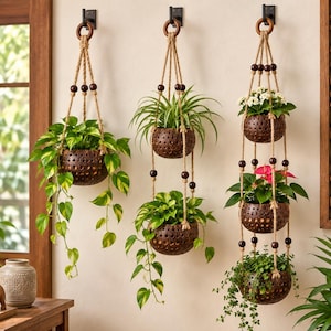 May include: Three hanging planters made from dark brown coconut shells with perforated patterns. The planters are suspended by thick jute ropes and dark wooden beads, featuring a single-tier, two-tier, and three-tier arrangement with green plants.