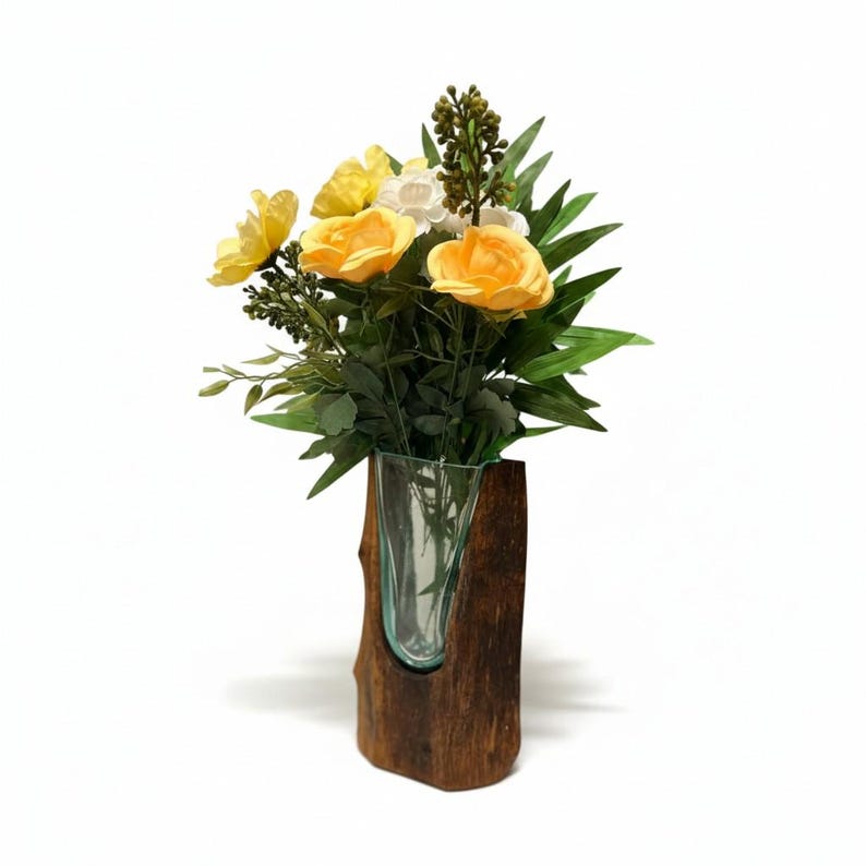 May include: A glass vase with a wooden base holds a bouquet of yellow and white flowers with green leaves. The vase is clear, and the wooden base is a dark brown color.