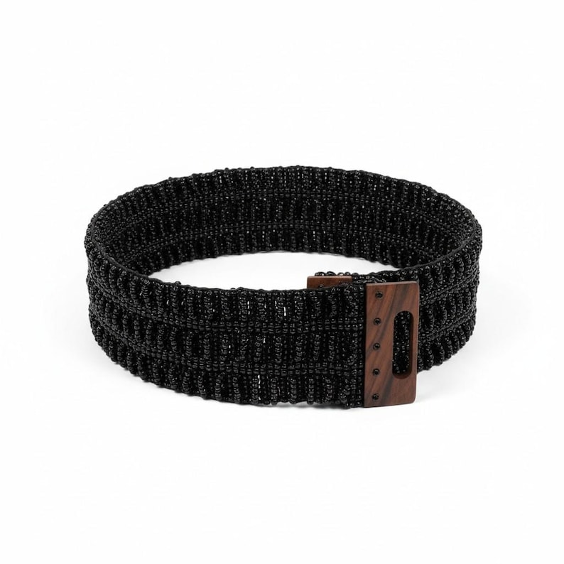 Pode incluir: Um cinto preto tecido com uma fivela retangular de madeira. O cinto &eacute; feito de pequenas contas pretas bem tecidas. A fivela de madeira &eacute; de cor castanho escuro e tem pequenos rebites decorativos.