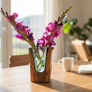 May include: A wooden and glass vase holds vibrant purple and pink gladiolus flowers. The vase sits on a light-colored wooden table, next to a white mug and an open book. The scene is bathed in natural light.