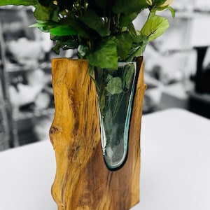 May include: A wooden vase with a clear glass insert holding white roses and green leaves. The vase is made of a light brown, textured wood, and the glass insert is a curved shape. The background is blurred, with a black and white color scheme.