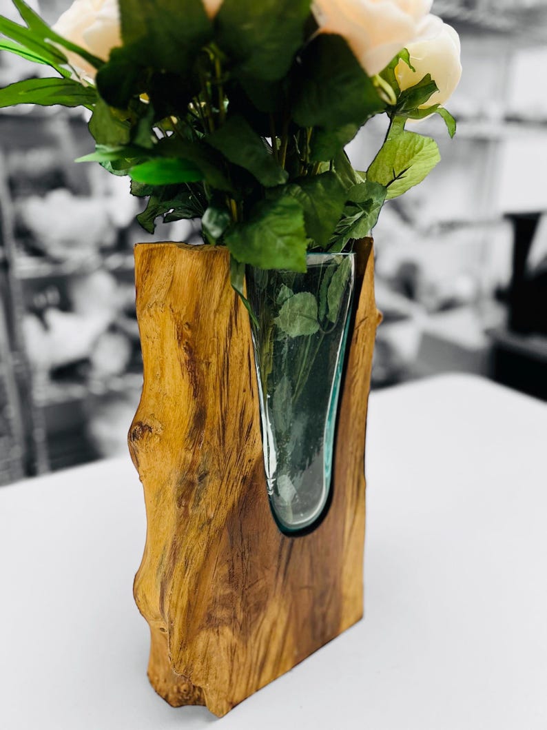 May include: A wooden vase with a clear glass insert holding white roses and green leaves. The vase is made of a light brown, textured wood, and the glass insert is a curved shape. The background is blurred, with a black and white color scheme.