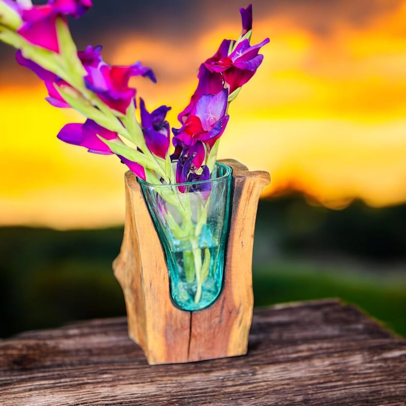 May include: A wooden vase with a teal glass insert holds vibrant purple and pink gladiolus flowers. The vase has a natural, rustic look, with the wood grain visible. The background features a blurred sunset with orange and yellow hues.