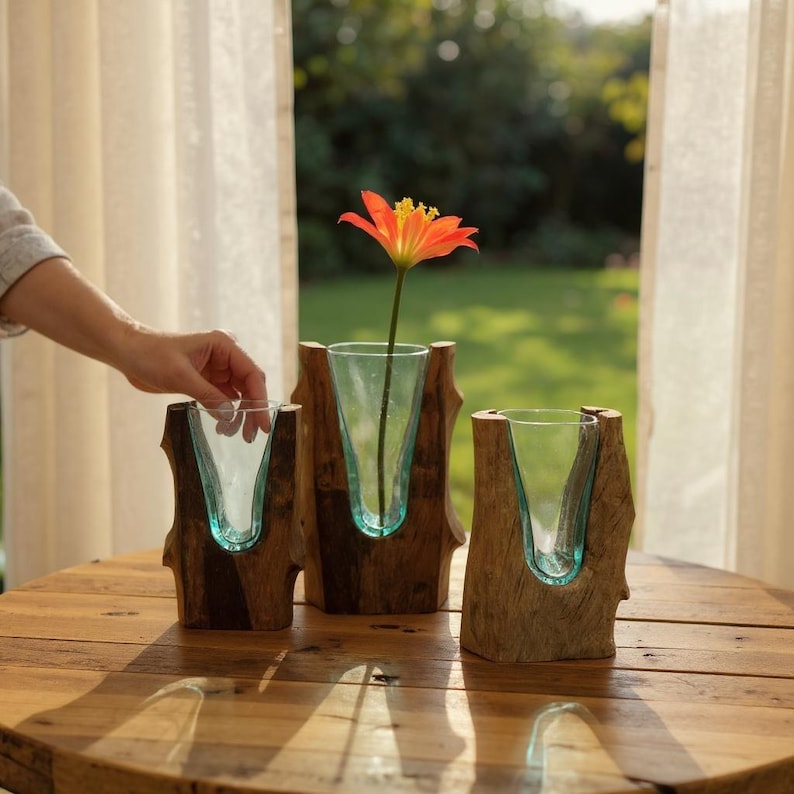 May include: Three wooden vases with clear glass inserts. One vase holds an orange flower with a yellow center. The vases are on a wooden table, with a hand reaching towards one. The background is a blurred view of a green yard.